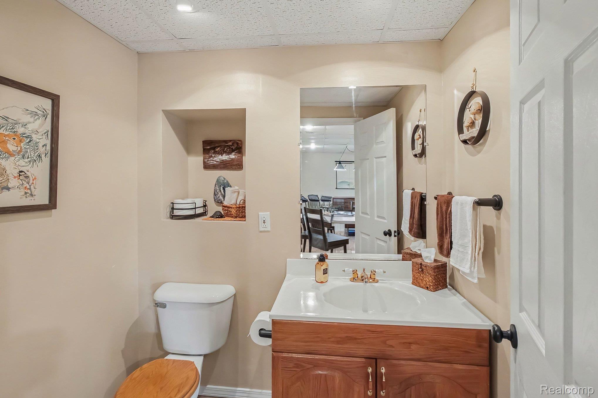 Bathroom with a wood vanity, white sink, toilet, and mirror reflecting a dining area.