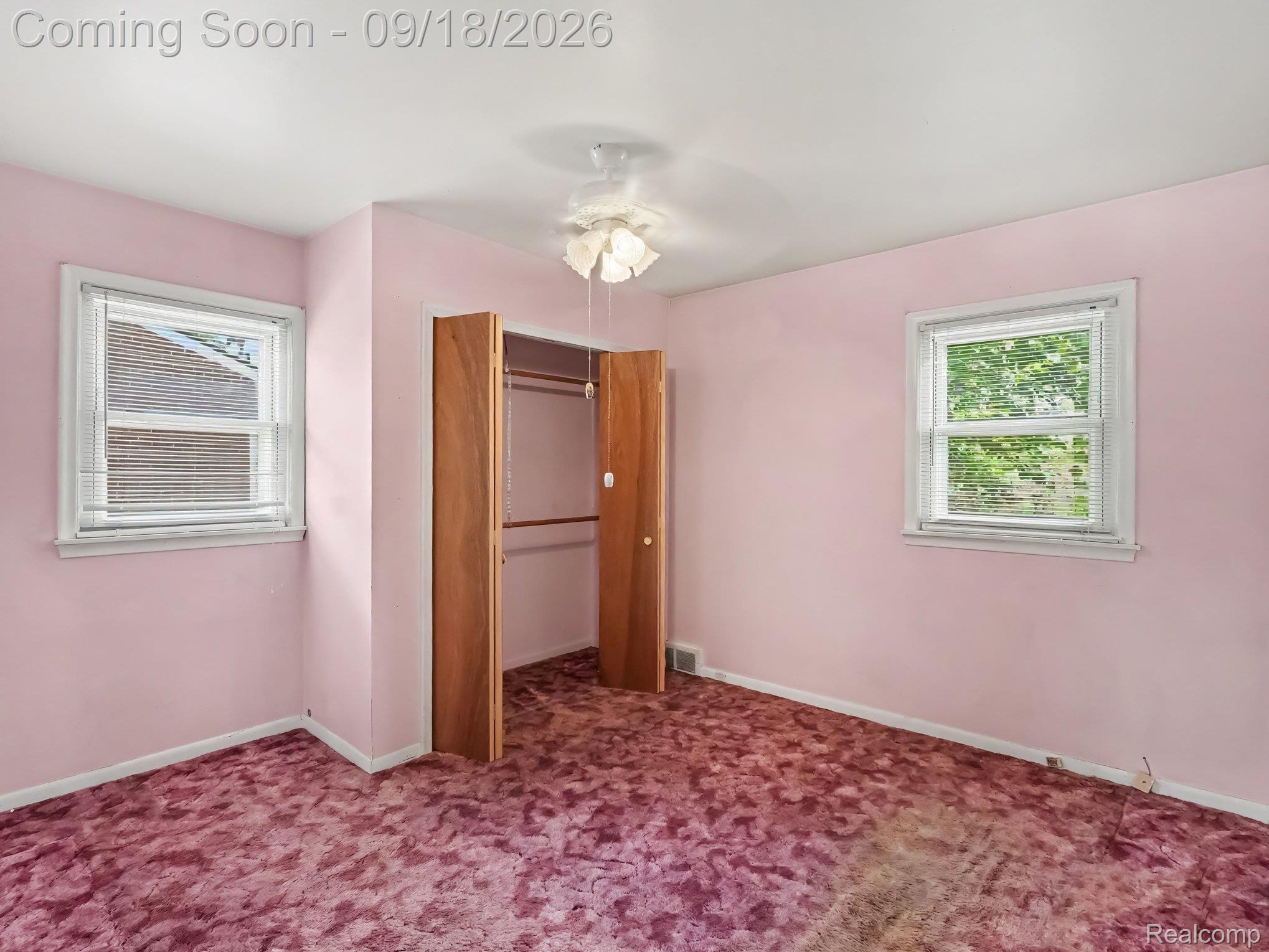 Empty bedroom with pink walls, patterned mauve carpet, two windows, and an open wooden closet.