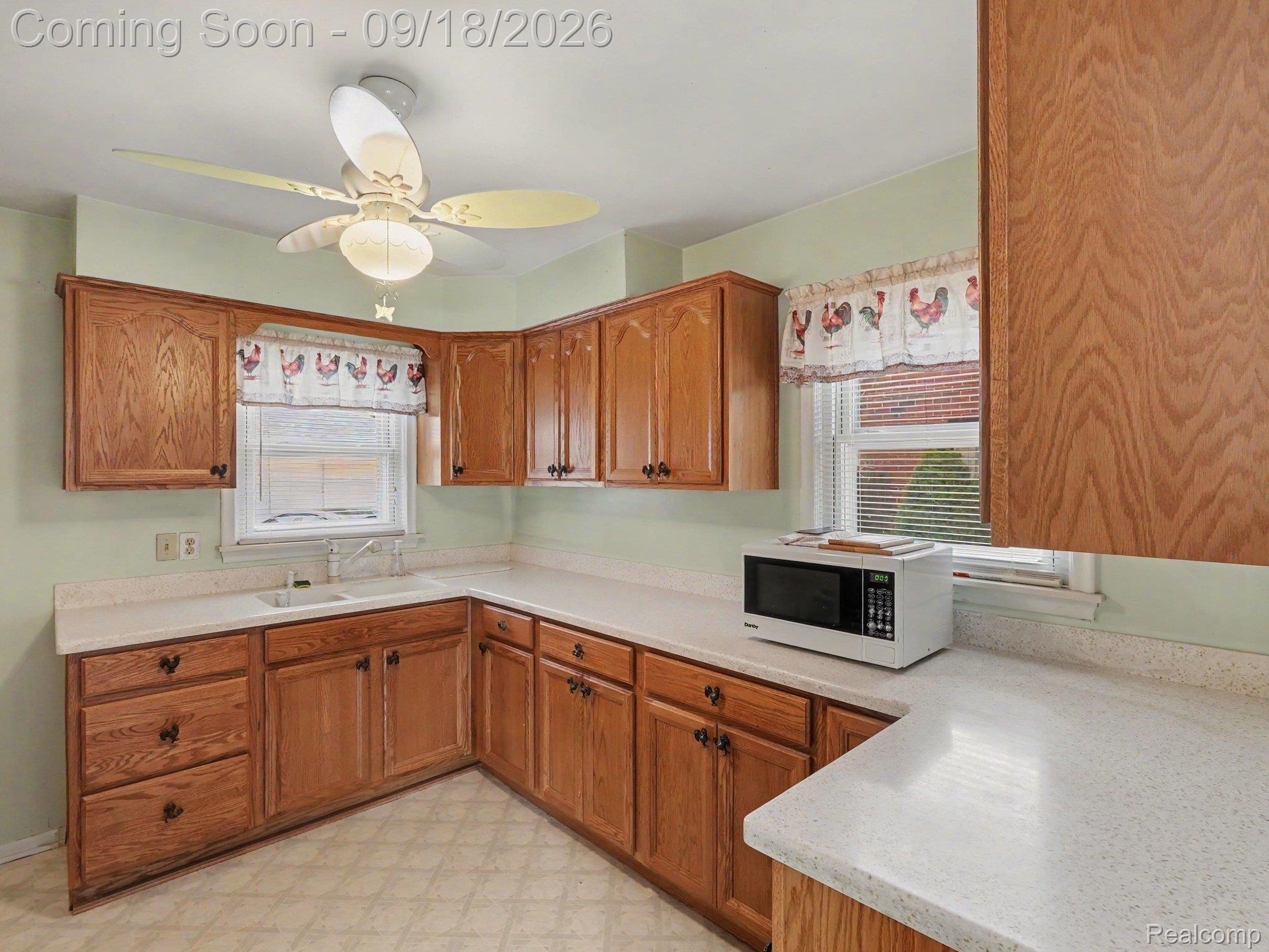 Kitchen with oak cabinets, pale countertops, a corner sink, and rooster-print window valances.