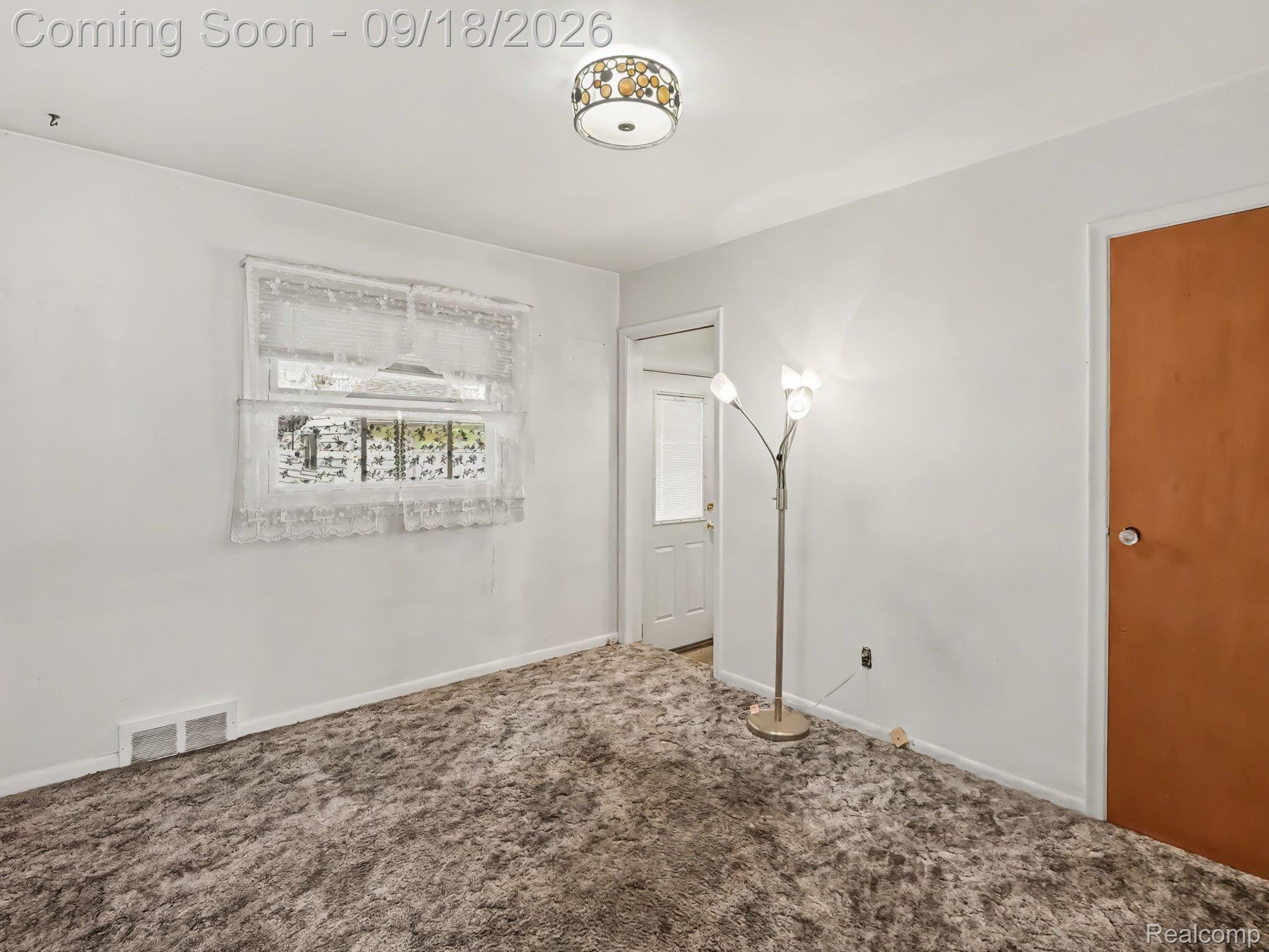 Mostly empty room with patterned carpet, a lace-curtained window, a floor lamp, and two doors.
