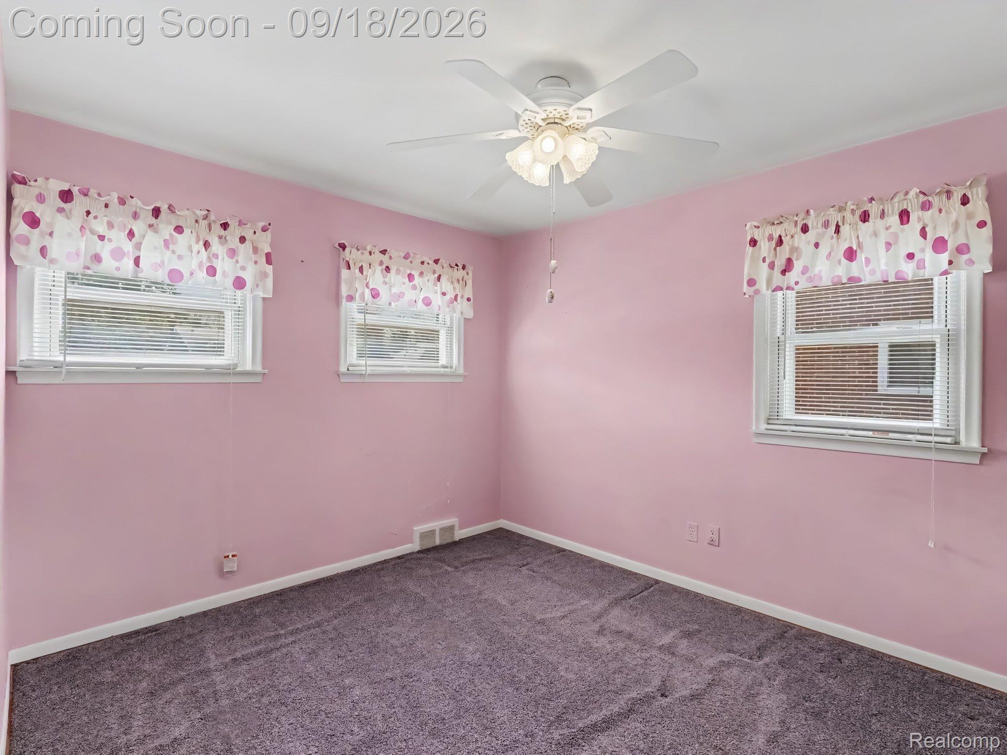 Empty pink bedroom with gray carpet, three curtained windows, and a ceiling fan.