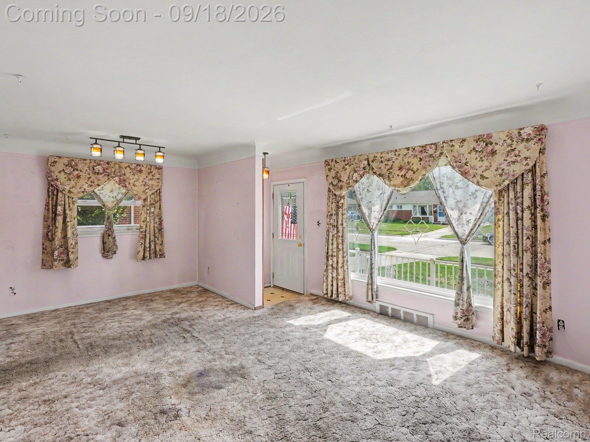 Empty living room with pale pink walls, floral curtains, and wall-to-wall carpet.