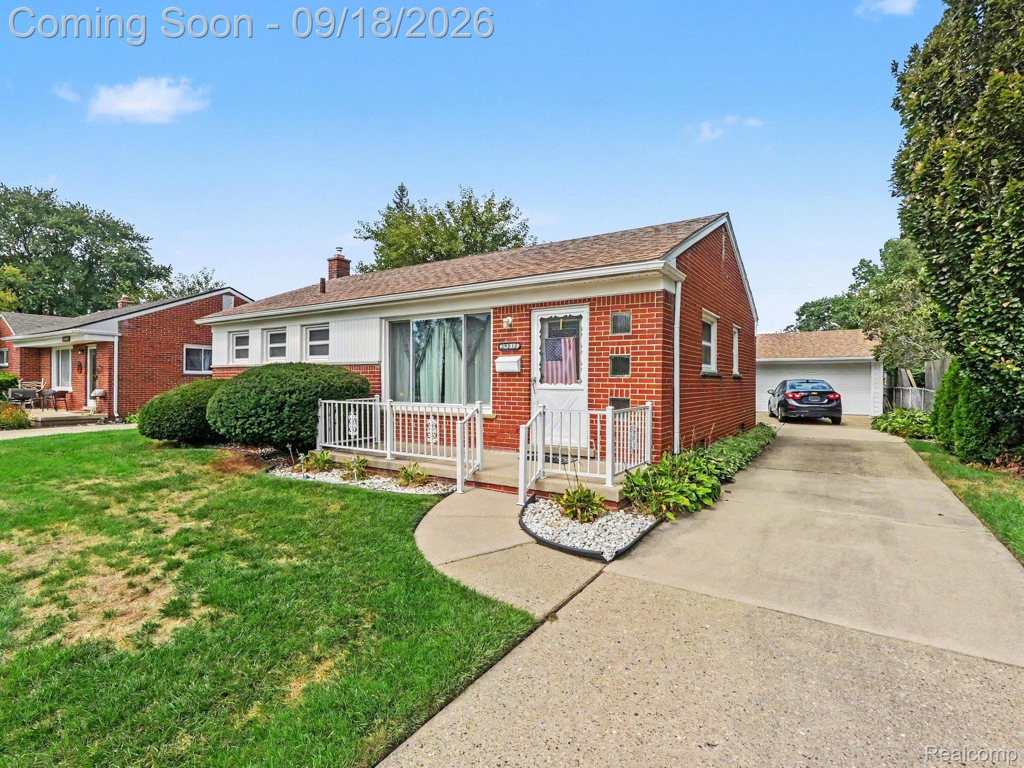 Red brick ranch house with a white-railed porch, green lawn, and driveway to a detached garage.