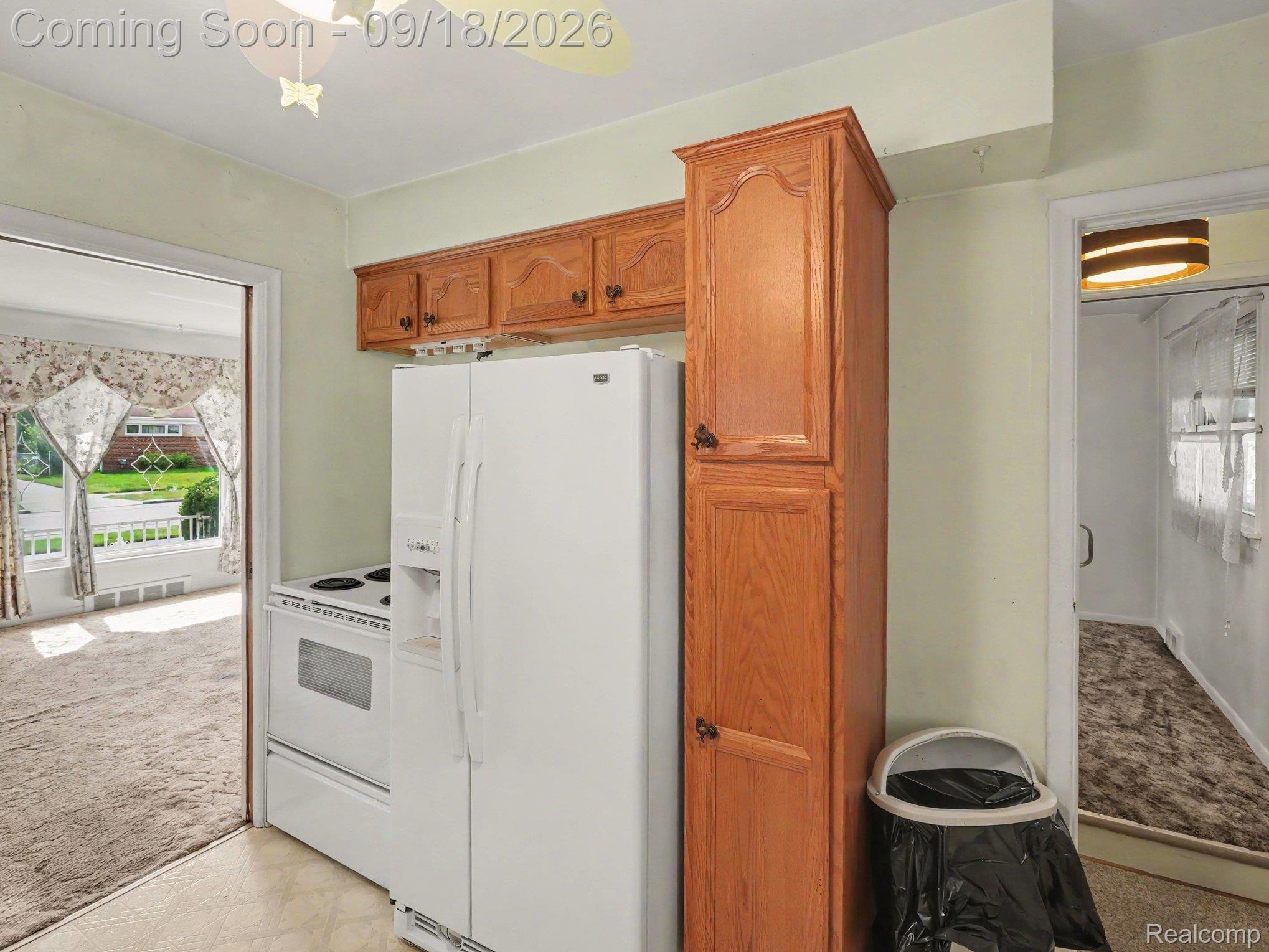 Kitchen with white appliances and oak cabinets, opening onto carpeted rooms.