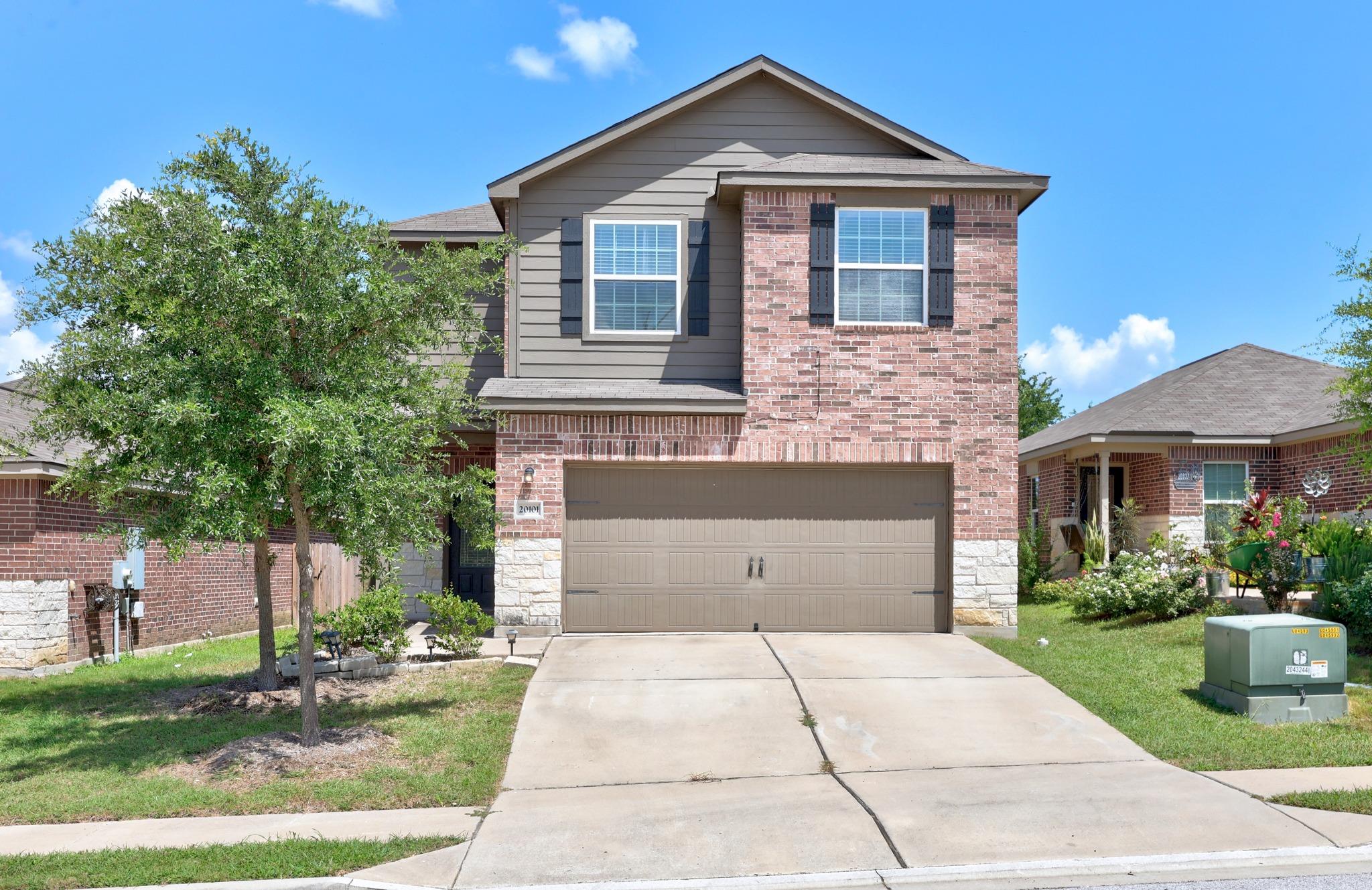 Two-story brick-and-gray house with a double garage, broad driveway, and mature tree in the front lawn.