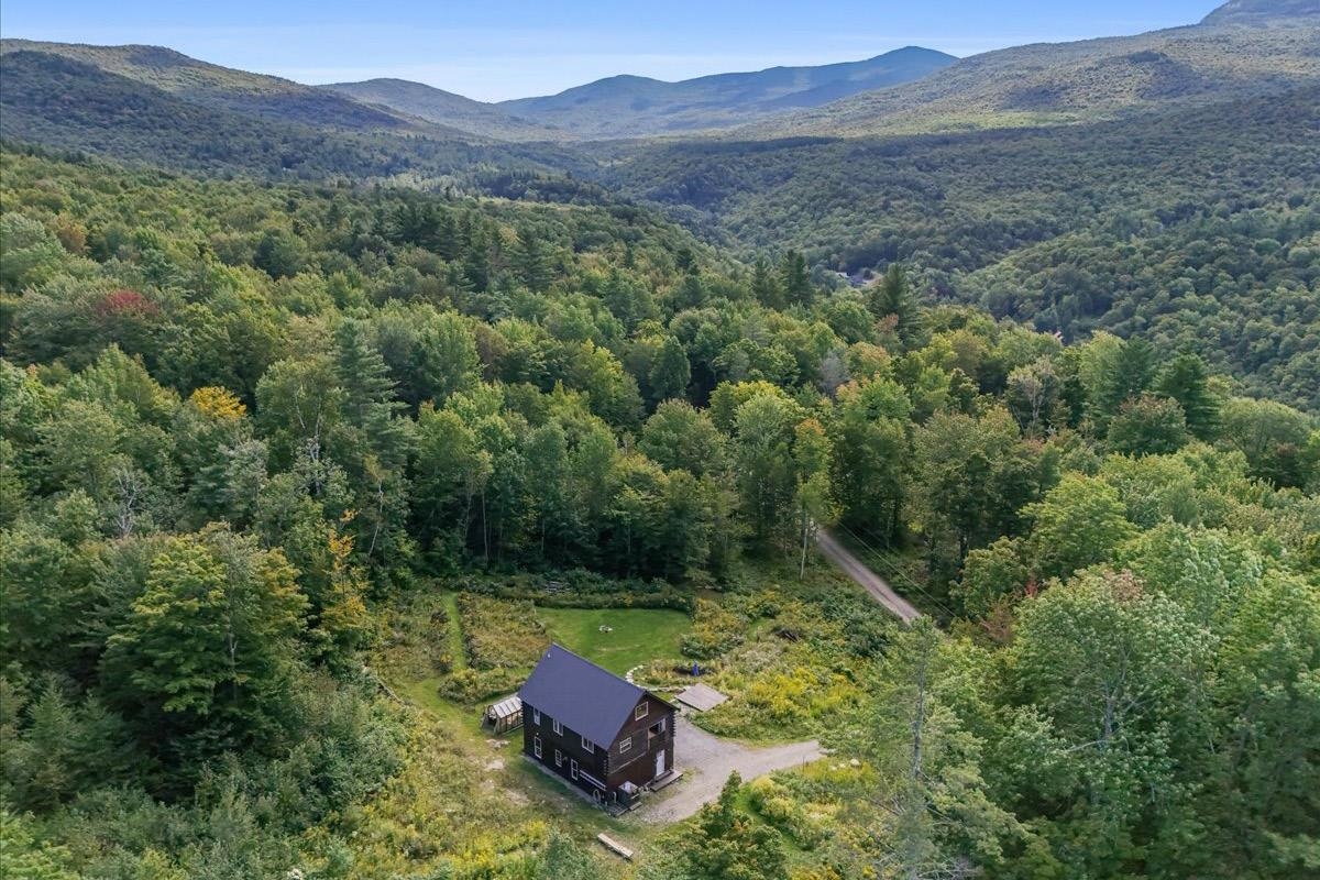 Aerial view of a two-story house in a forest clearing, with wooded mountains beyond.