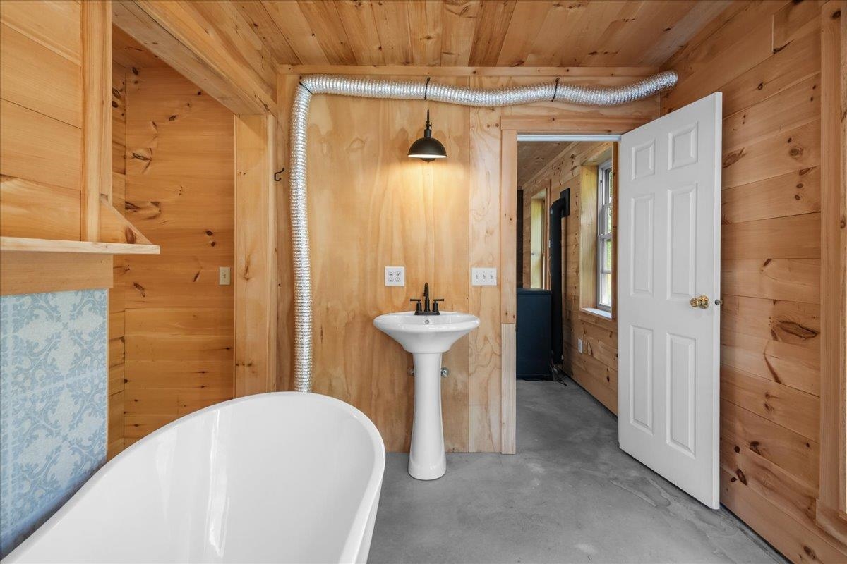 Wood-paneled bathroom with a freestanding tub, pedestal sink, and open door to an adjoining room.