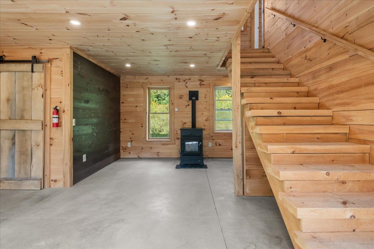 Wood-paneled interior with a wooden staircase, black wood stove, and concrete floor.
