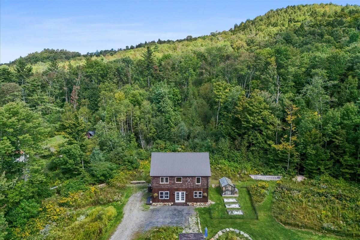 Aerial view of a rustic cabin with a metal roof amid a wooded hillside and garden beds.
