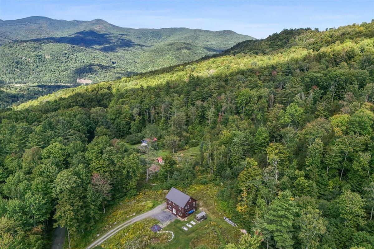 Aerial view of a mountain home surrounded by dense forest, with gardens and a winding driveway.