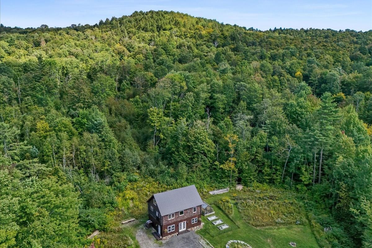 Aerial view of a two-story house beside a wooded hillside, with garden plots and a gravel driveway.