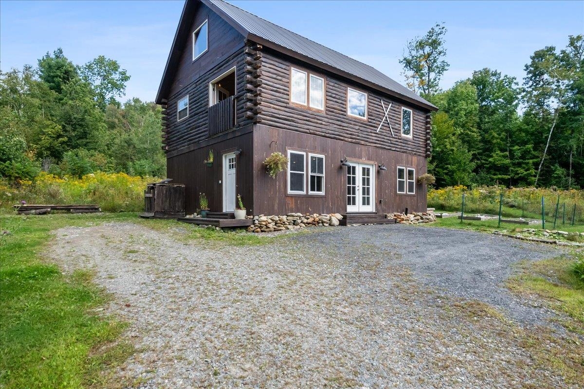 Two-story log home with a metal roof, small balcony, and gravel driveway surrounded by greenery.