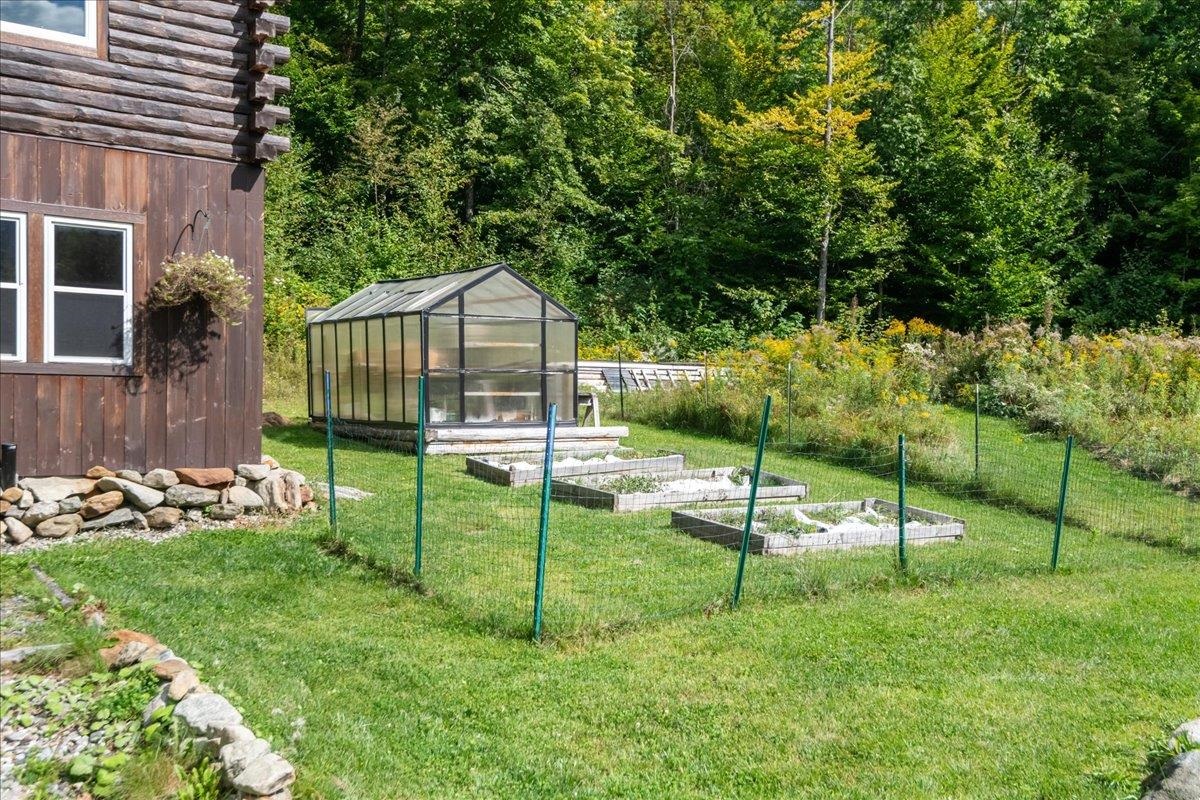 Greenhouse and raised garden beds beside a log-sided home, with woods in the background.