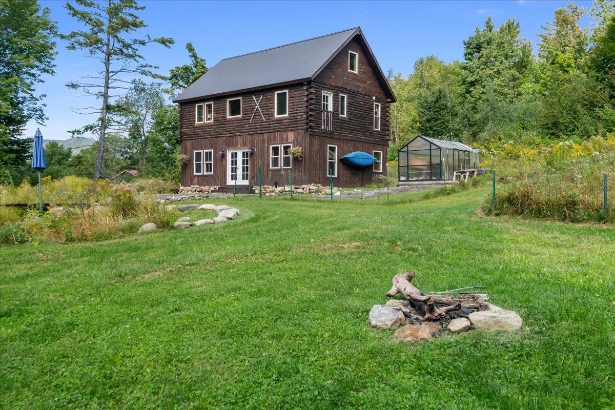 Two-story log home with a metal roof, greenhouse, and fire pit in a wooded lawn.