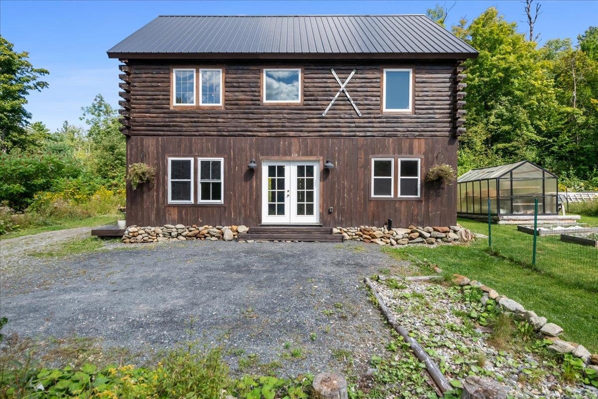 Two-story log house with a metal roof, gravel driveway, and greenhouse beside it.