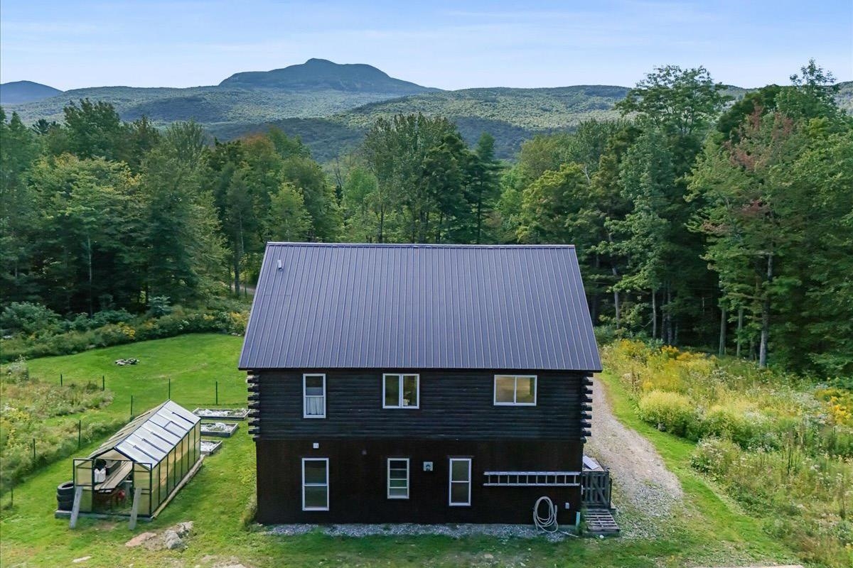 Dark log-sided home with a greenhouse in a wooded clearing, backed by mountain views.