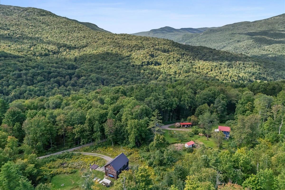 Aerial view of a rural home and outbuildings amid forested mountain slopes.
