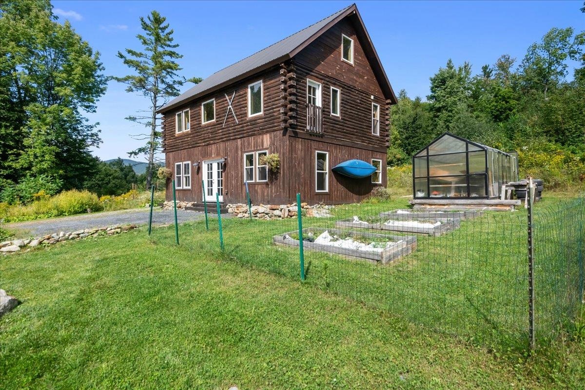 Two-story log home with a metal roof, greenhouse, raised garden beds, and fenced yard.