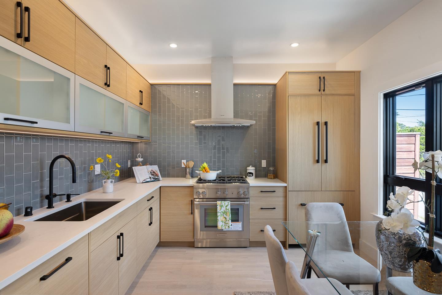 Kitchen with light wood cabinets, gray tile backsplash, stainless steel range, and glass dining table.