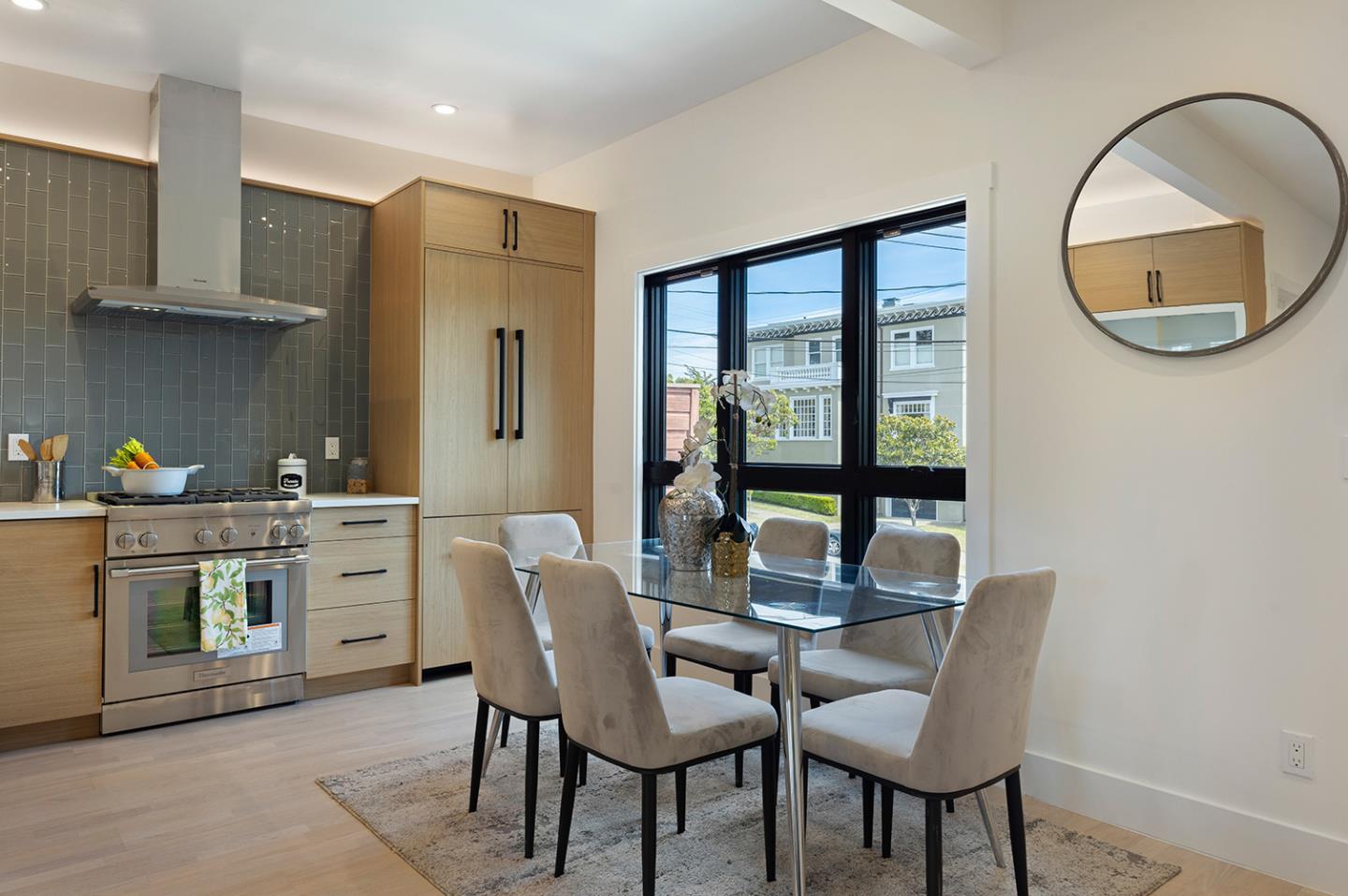 Kitchen and dining area with light wood cabinets, gray tile backsplash, and a glass dining table by large windows.