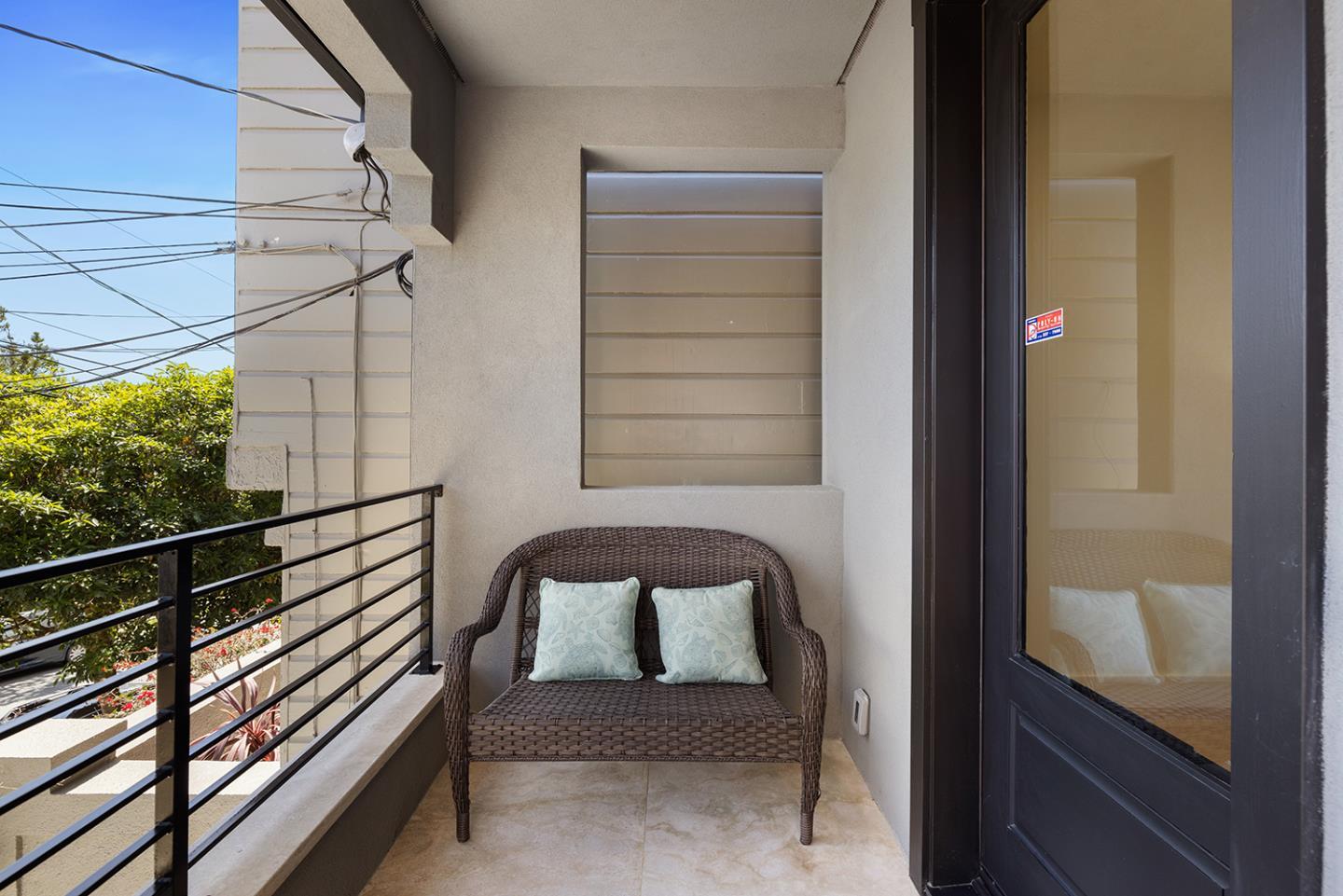 Balcony with a wicker chair, tiled floor, and black railing overlooking trees and rooftops.