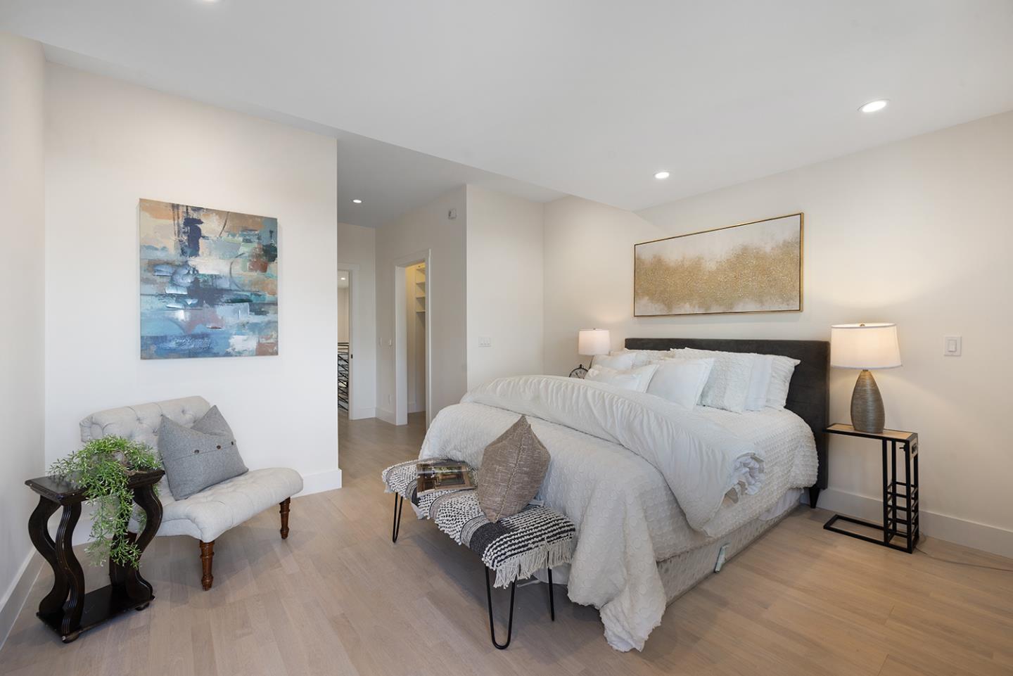 Staged bedroom with white bedding, abstract wall art, a bench at the foot of the bed, and a corner chair.