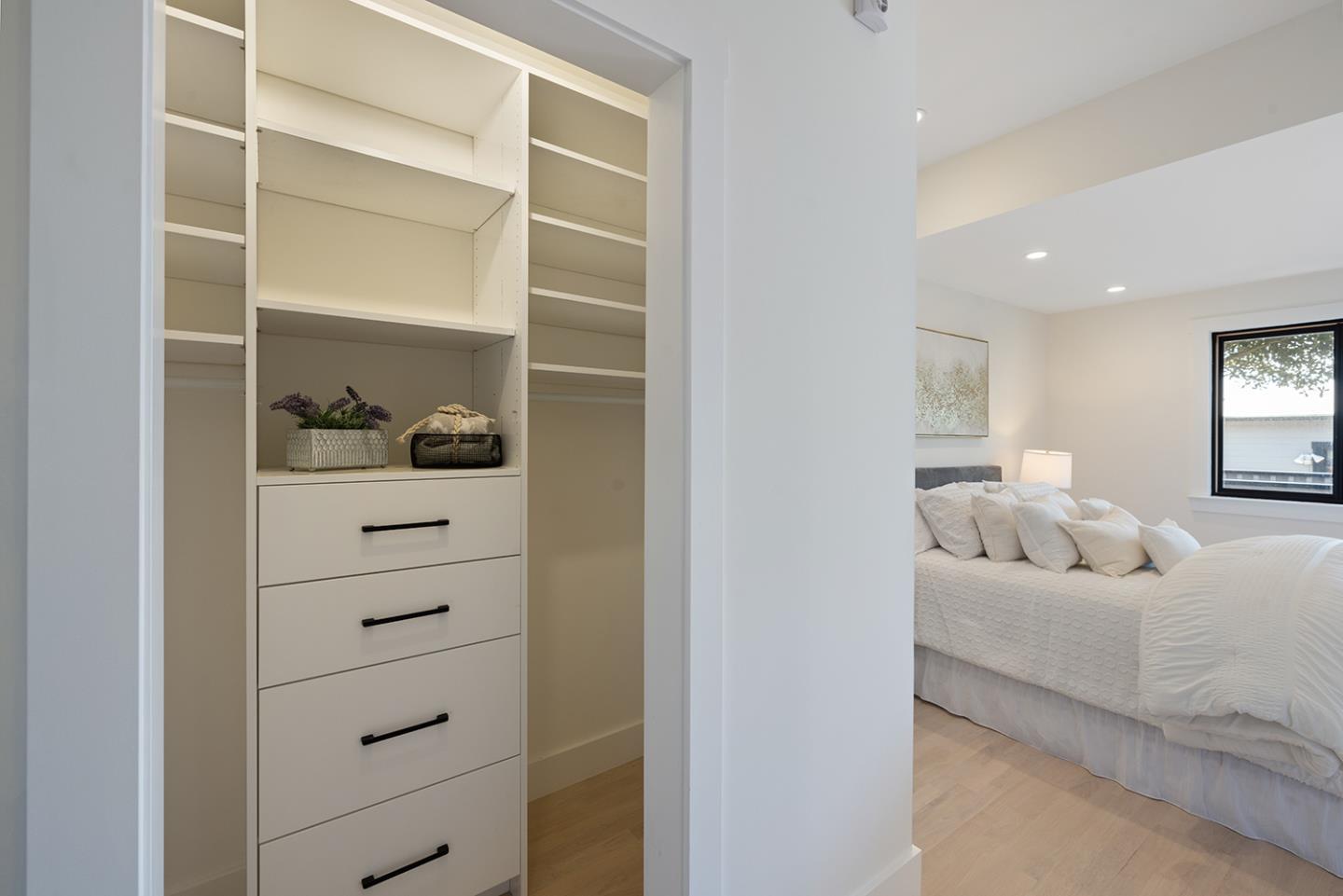 Staged bedroom with a white bed beside a built-in closet with open shelves and drawers.