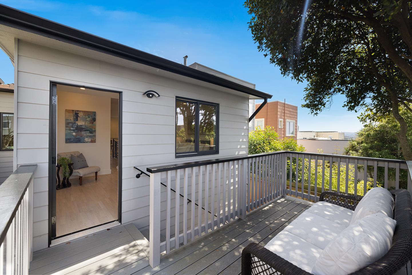 Furnished deck with a white-cushioned wicker sofa, gray decking, and an open doorway into the home.