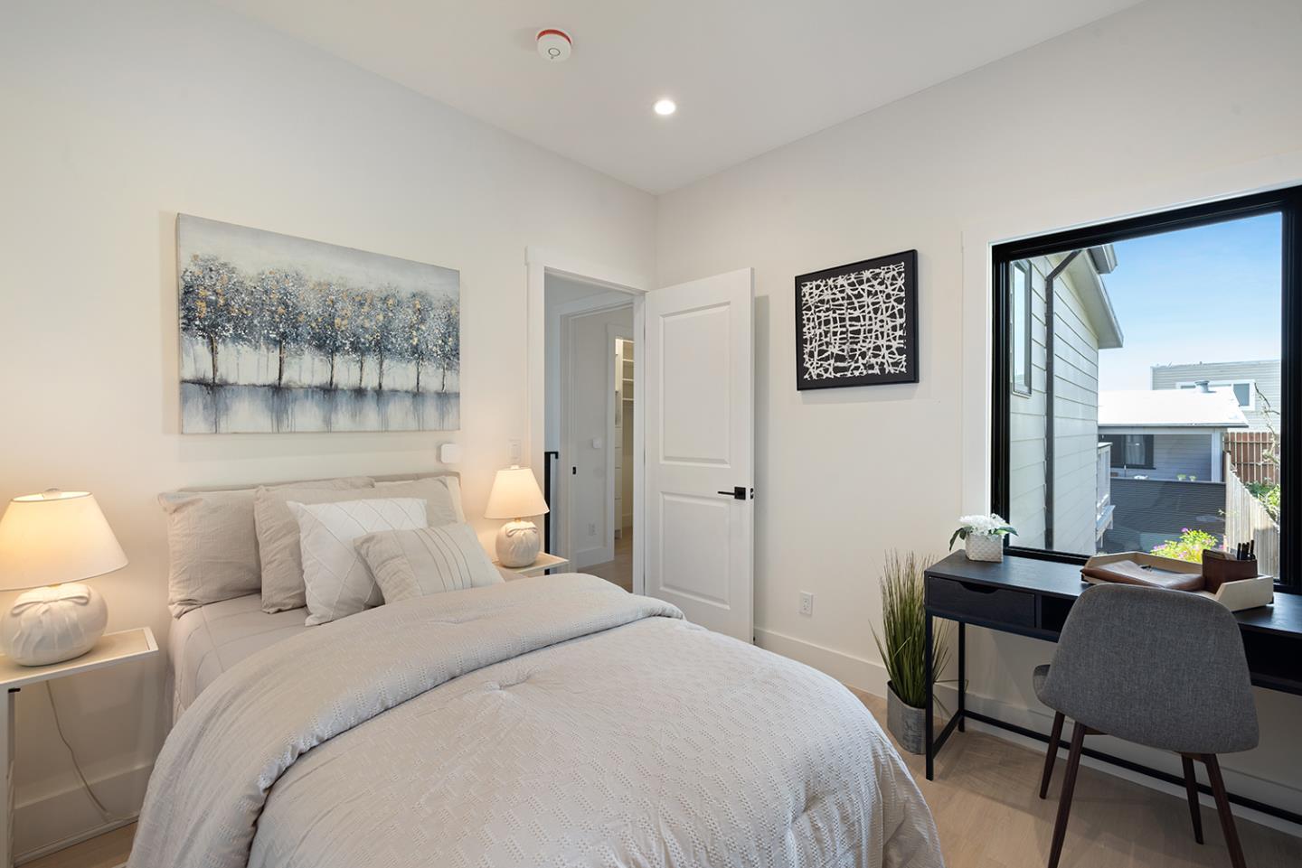 Staged bedroom with a white bed, abstract wall art, and a desk beside a large window.