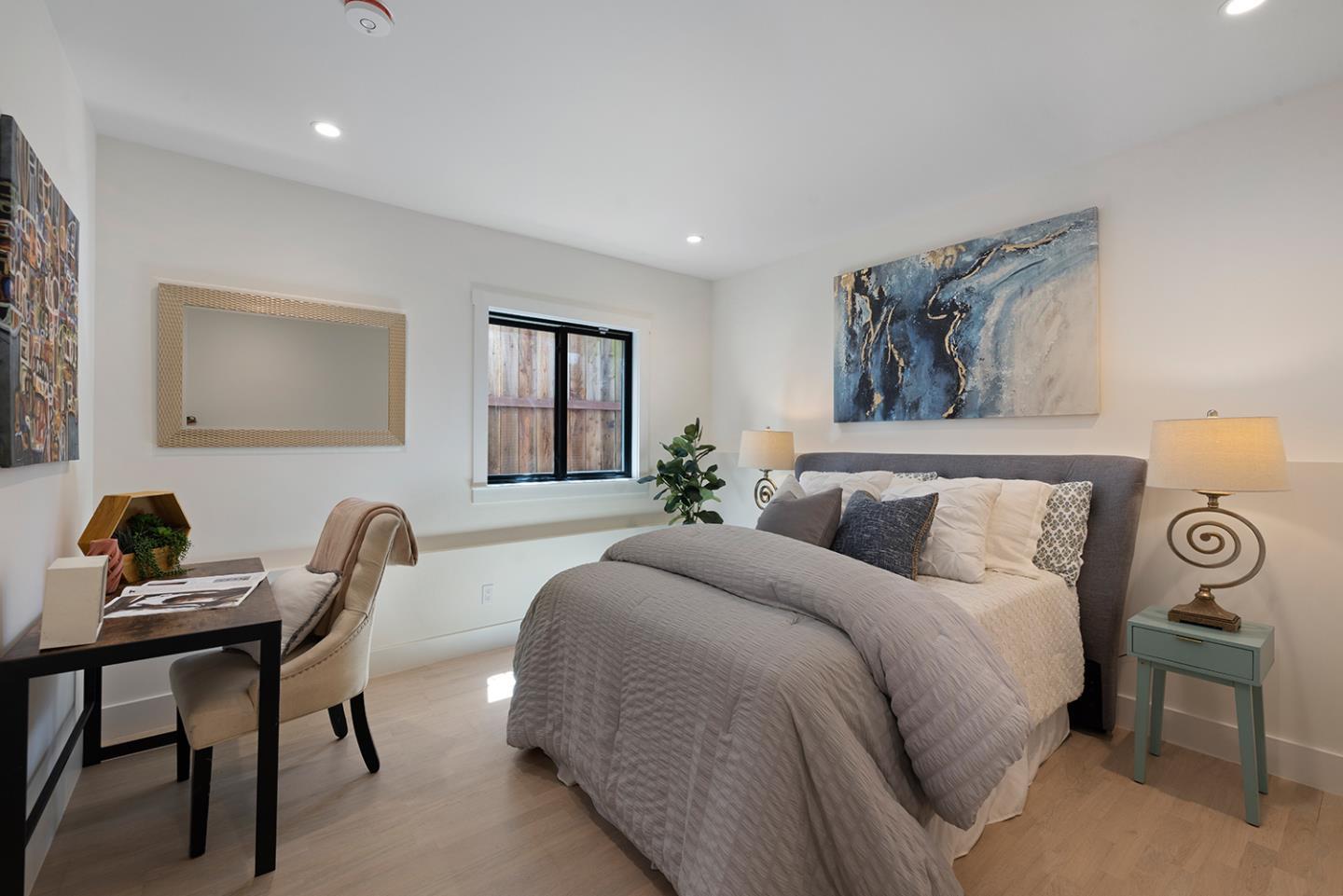 Staged bedroom with a gray upholstered bed, desk, and window overlooking a wooden fence.