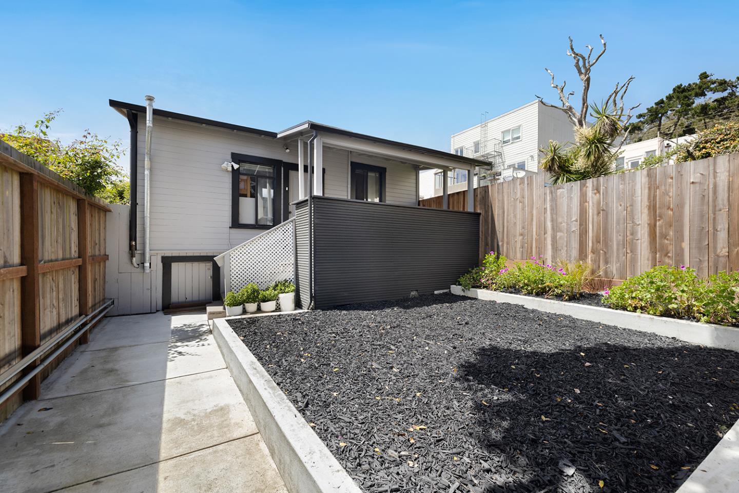 Rear of a gray-sided house with a raised deck, fenced yard, and large mulched garden bed.