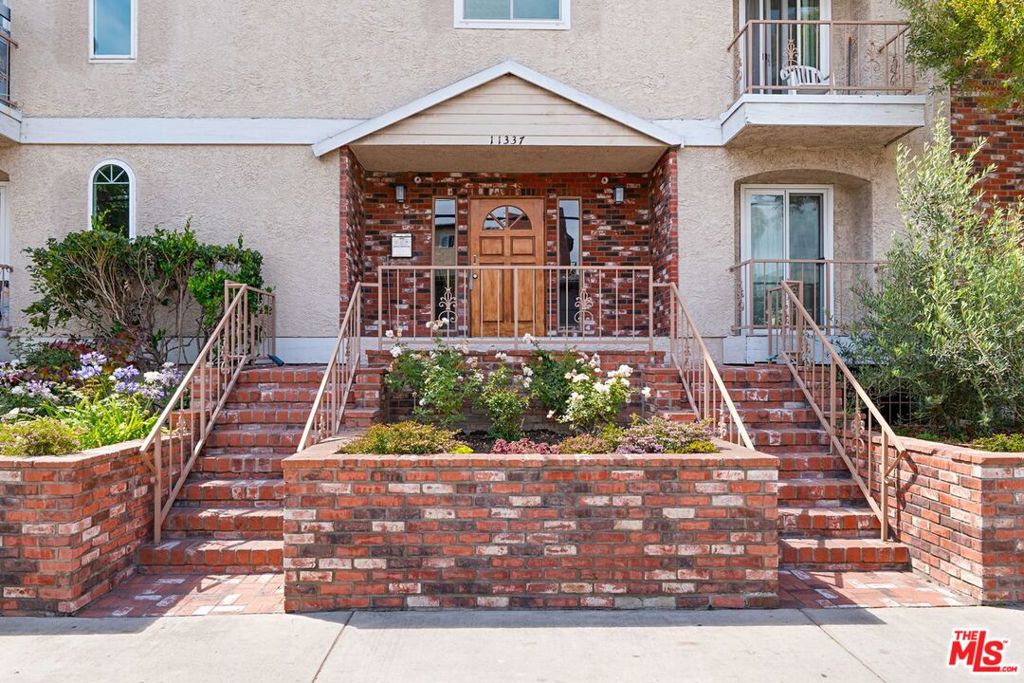 Residential building entrance with brick stairs, metal railings, and landscaped flower beds.