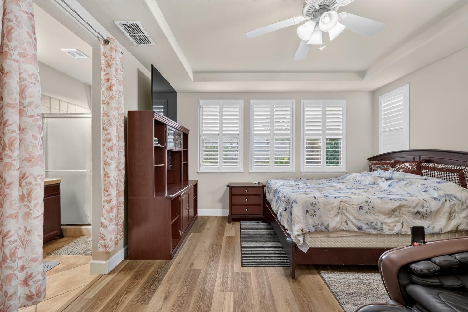 Bedroom with plantation shutters, a tray ceiling, and dark wood furniture beside a curtained bathroom doorway.