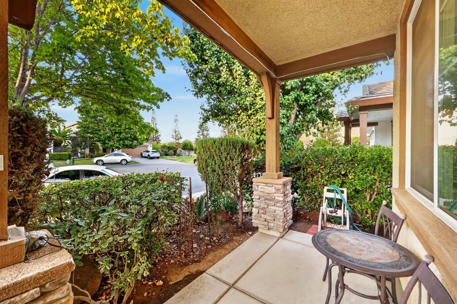 Covered patio with stone column, outdoor table, chairs, and leafy hedges overlooking a residential street.