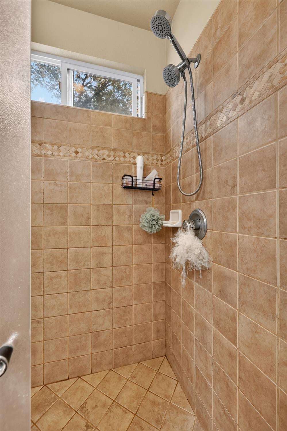 Tan tiled walk-in shower with a decorative border, dual showerheads, and a high window.