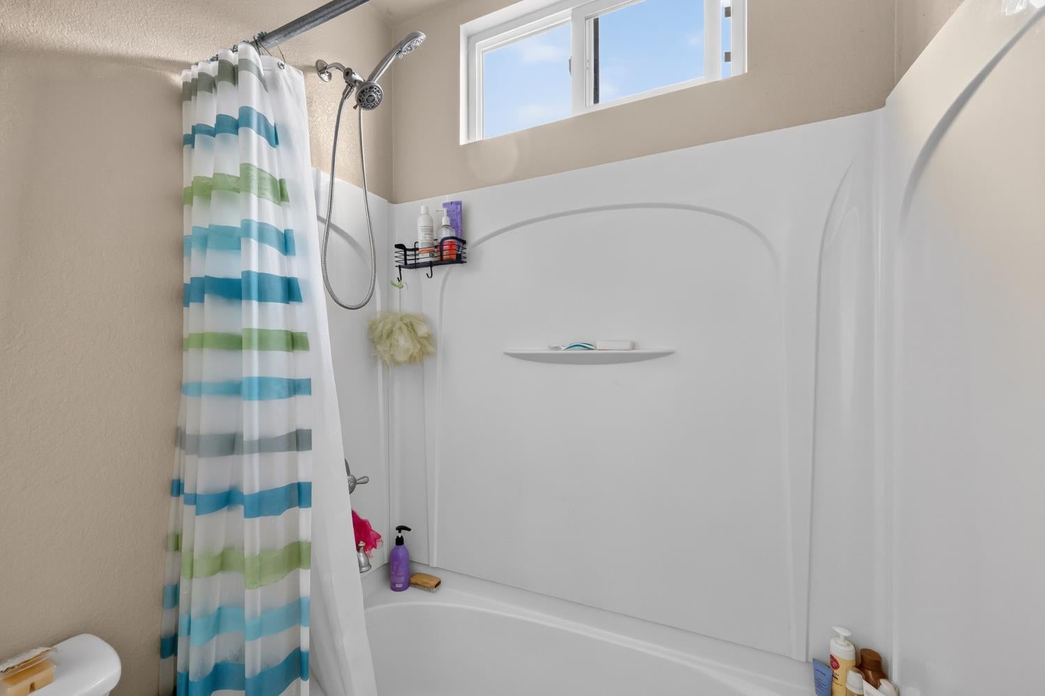 Bathroom with a white tub and shower, striped curtain, handheld showerhead, and high window.