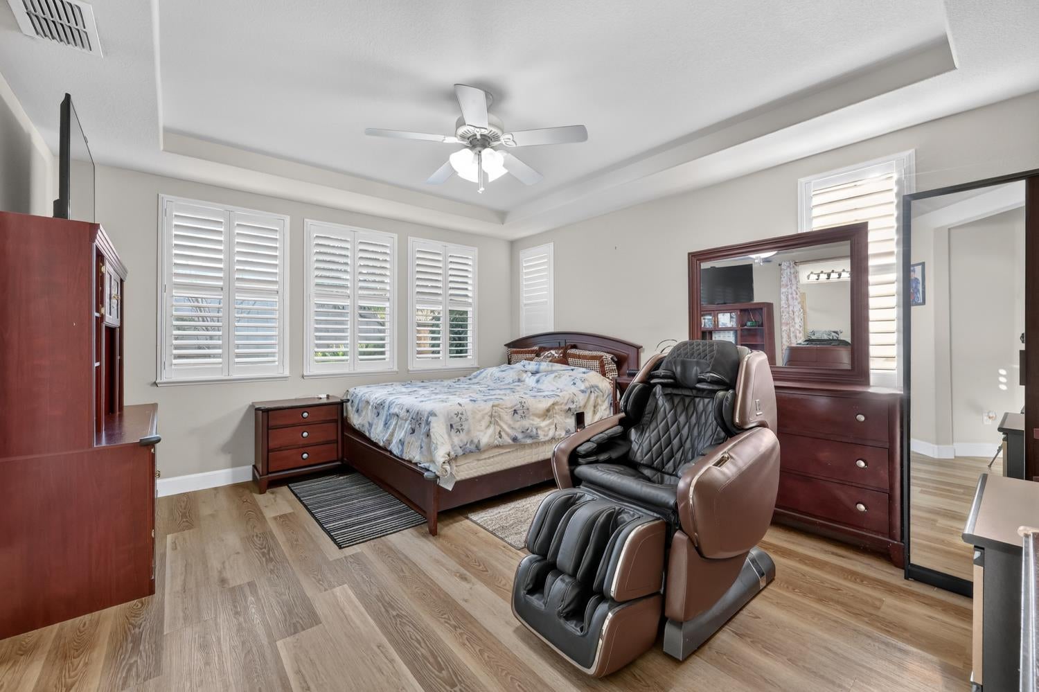 Bedroom with plantation shutters, tray ceiling, and a large massage chair beside the bed.