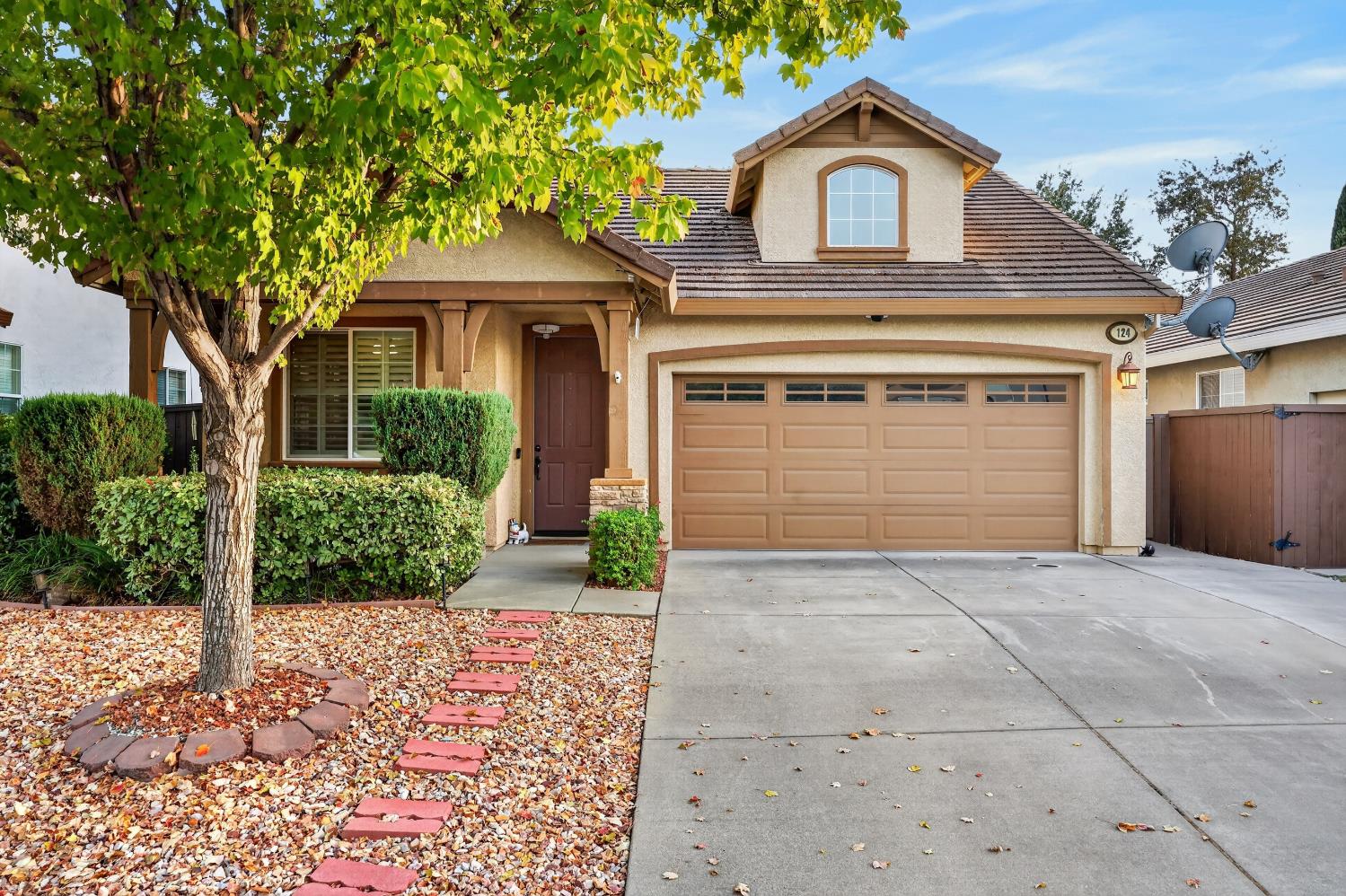 Two-story beige house with a wide garage, shaded entry, and leafy front yard.