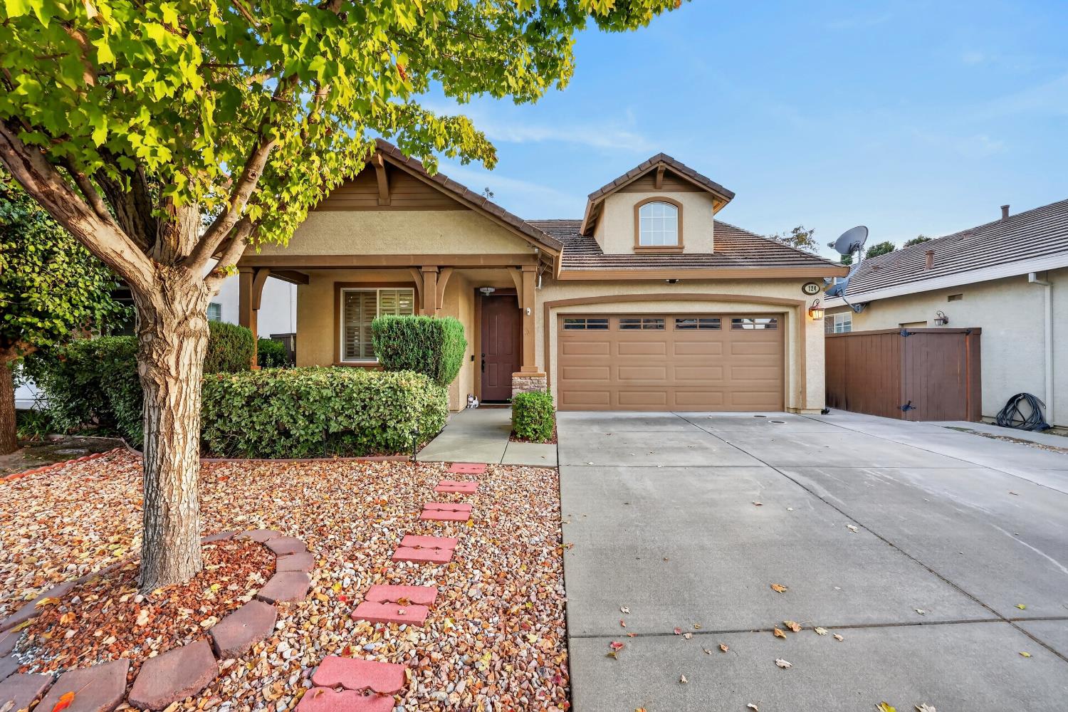 Tan stucco home with a two-car garage, broad driveway, and a large tree shading the landscaped front yard.