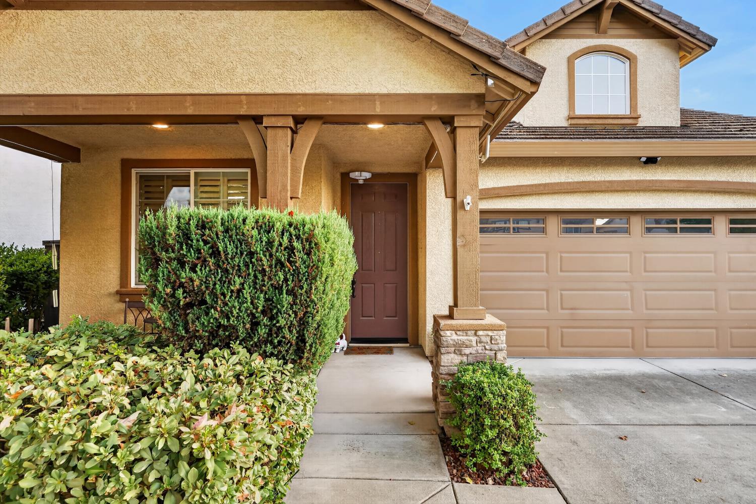 Two-story tan stucco house with a covered entry, brown garage door, and trimmed front shrubs.
