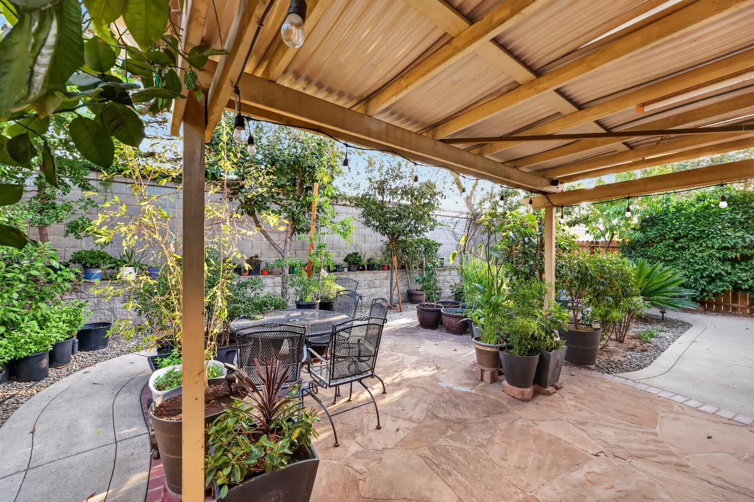 Covered backyard patio with a timber roof, stone paving, metal chairs, and abundant potted plants.