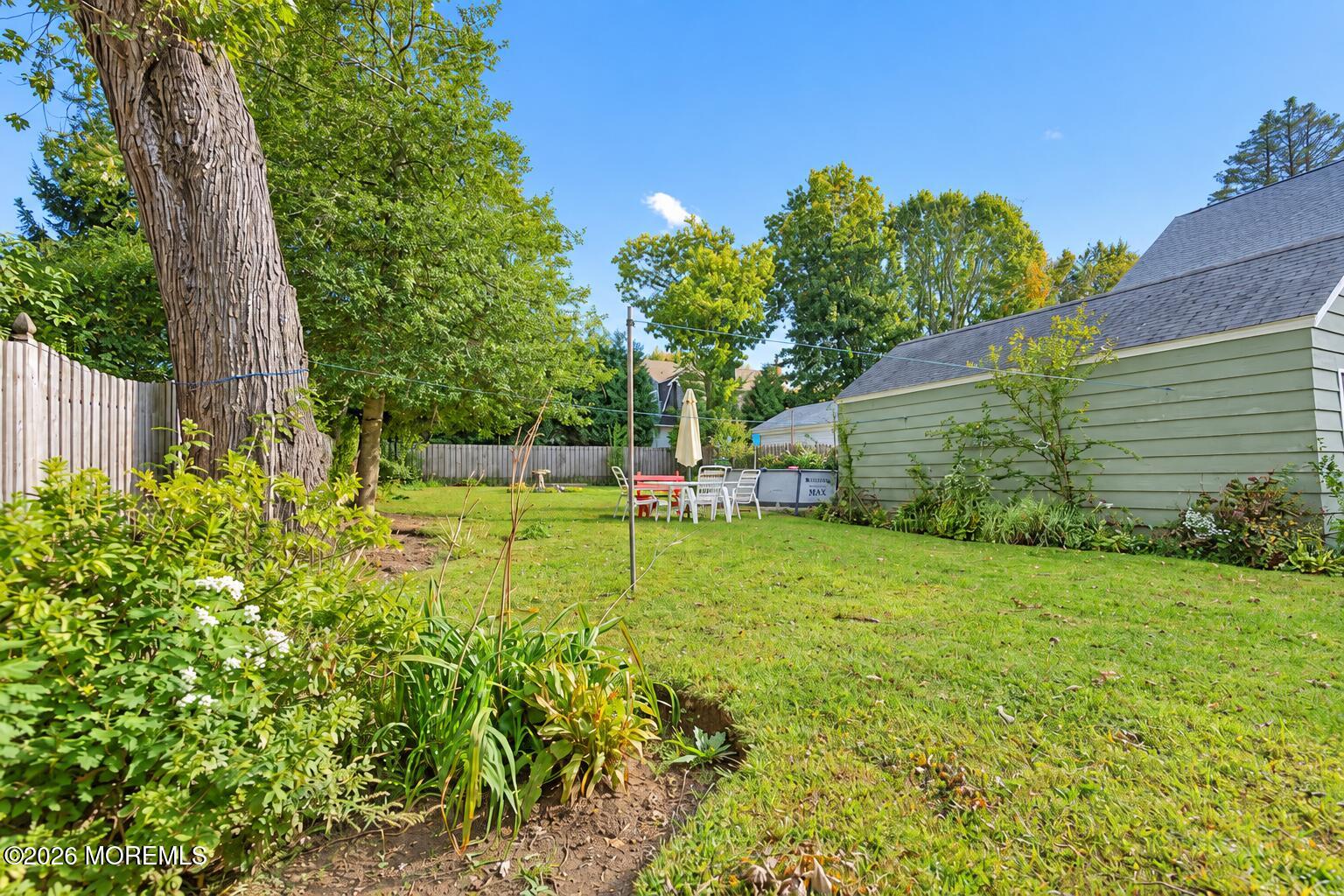 Backyard lawn with mature trees, garden beds, and outdoor dining furniture beside a light-green outbuilding.