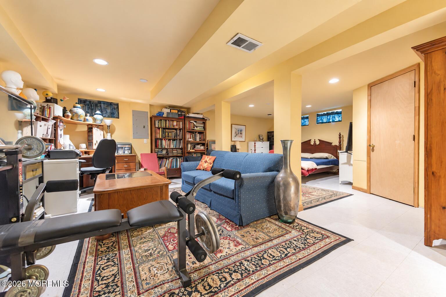 Furnished basement with a home office, exercise equipment, blue sofa, and adjoining bedroom.