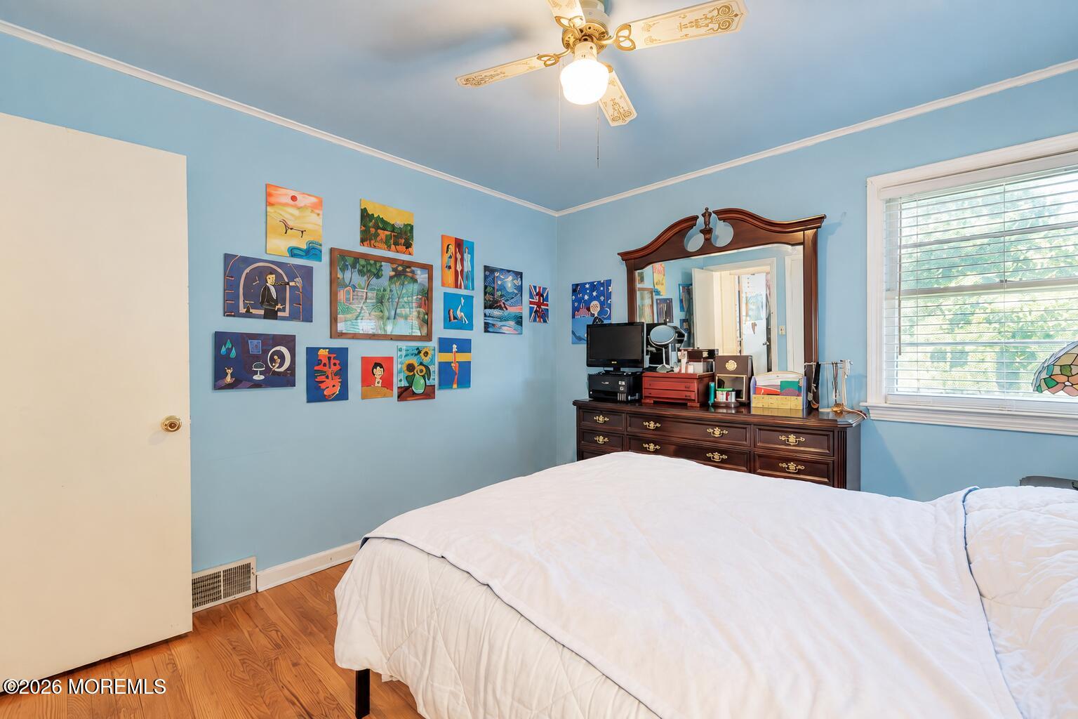 Bedroom with blue walls, colorful artwork, a dark wood dresser, and a white bed.