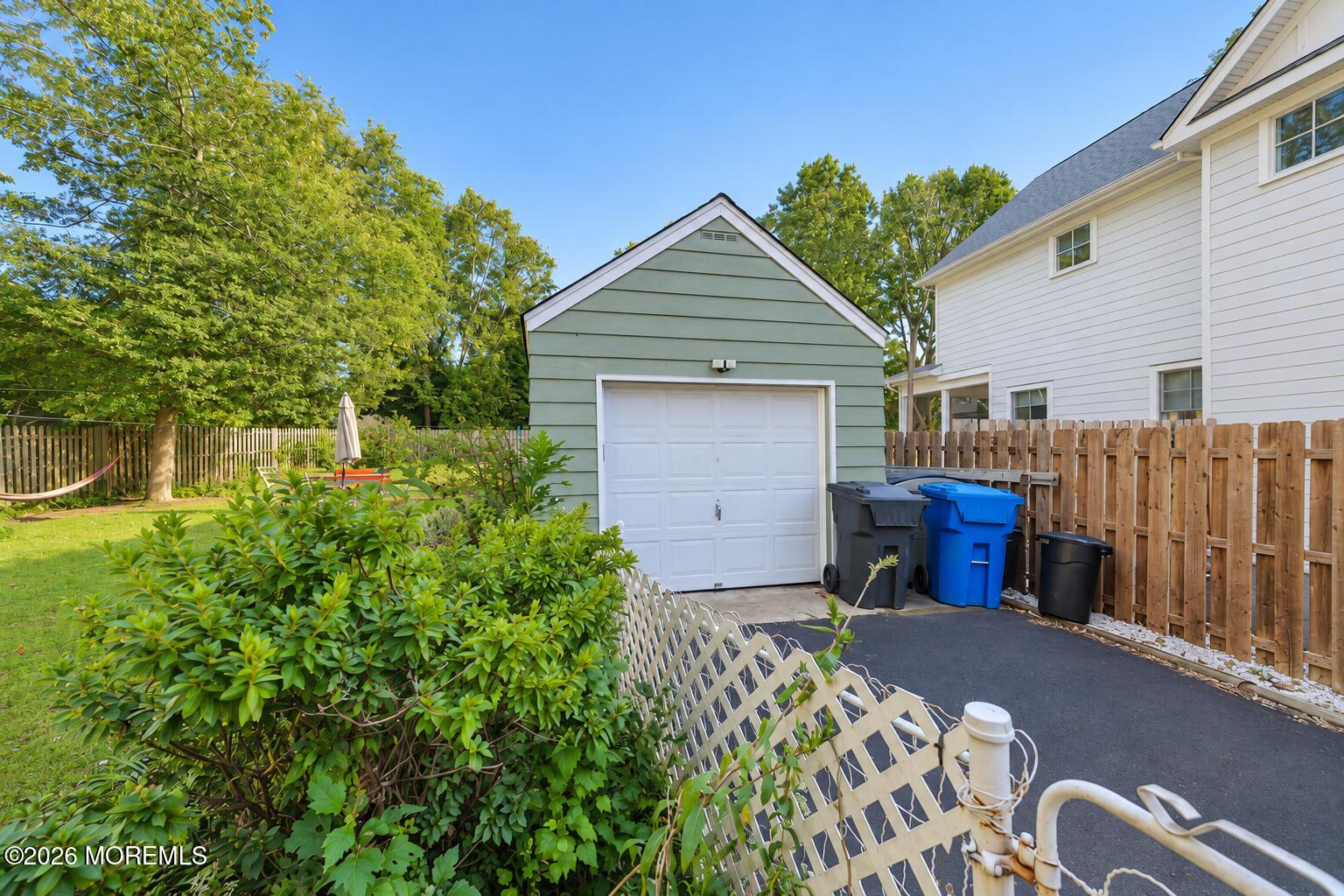 Detached garage beside a white house, with a fenced driveway and leafy backyard.
