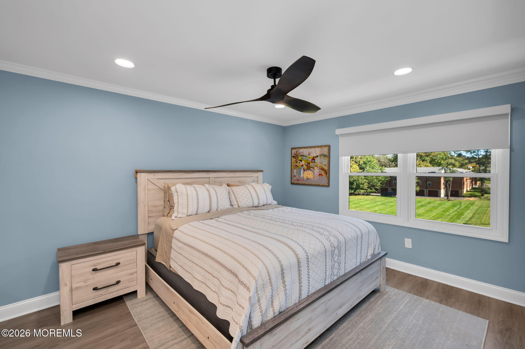 Furnished bedroom with blue walls, a light wood bed, and a wide window overlooking a green lawn.