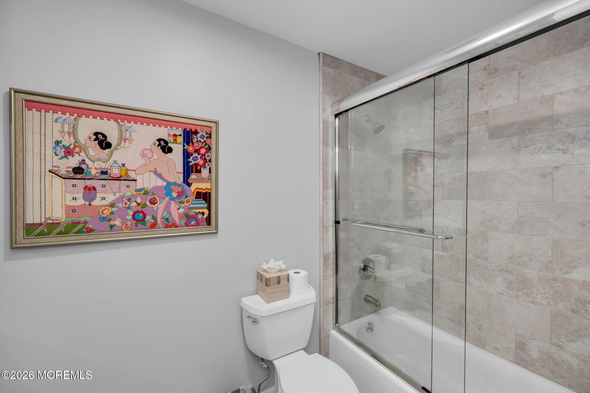 Bathroom with a glass-enclosed tub, beige tile surround, and colorful framed artwork.