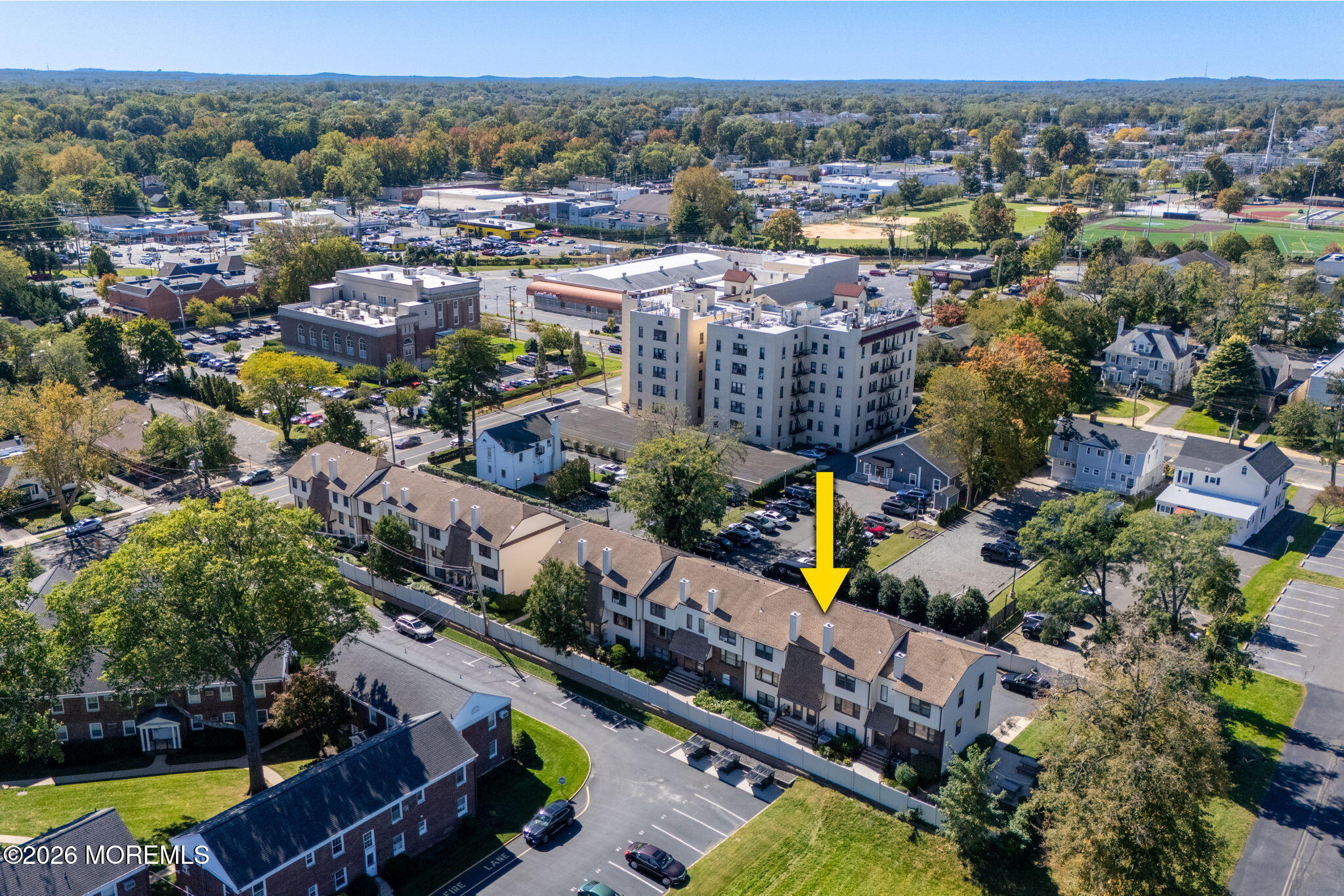 Aerial view of attached townhouses, with a yellow arrow marking one unit among nearby buildings and sports fields.