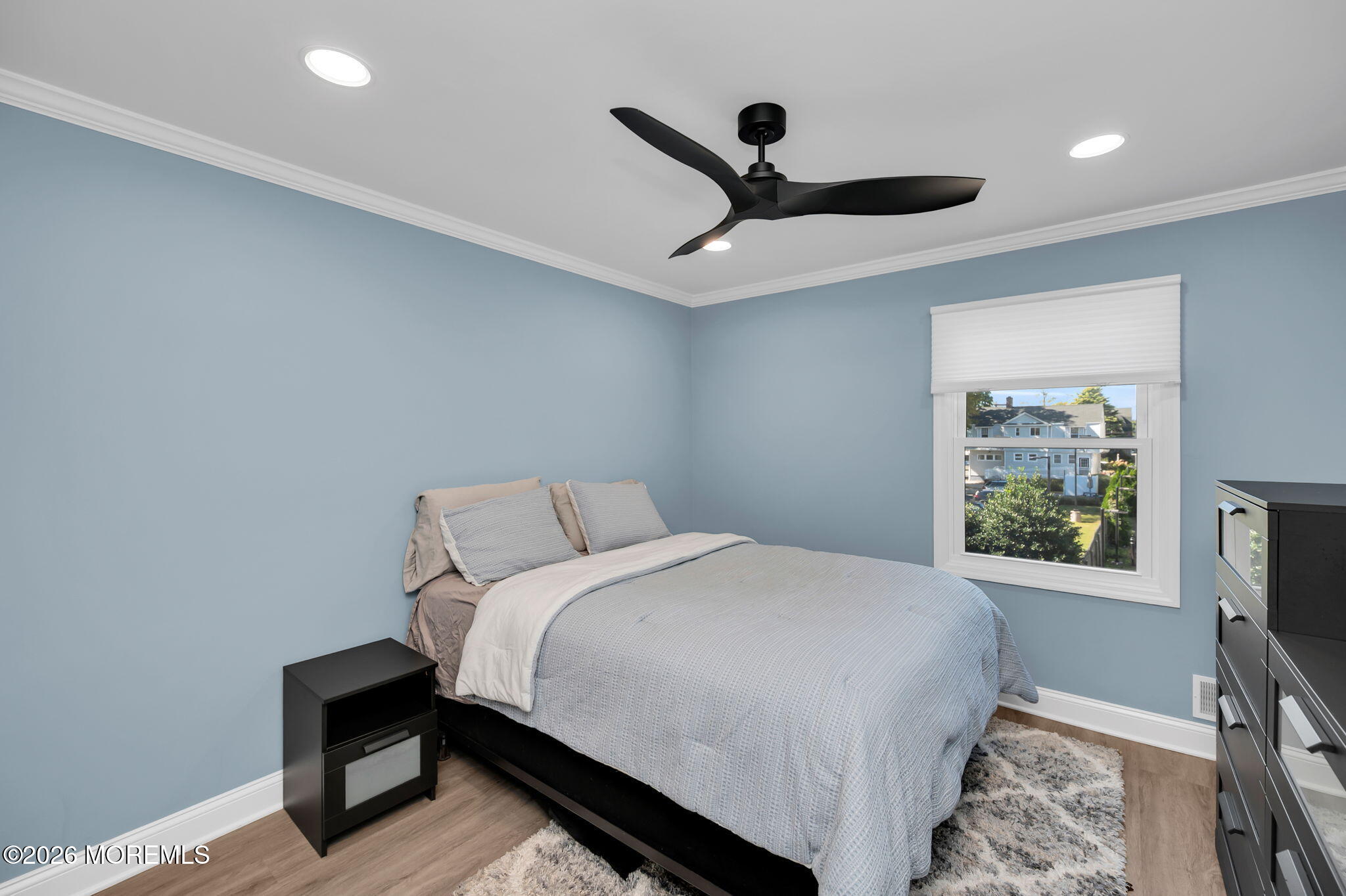 Bedroom with pale blue walls, light gray bedding, and a window overlooking neighboring homes.