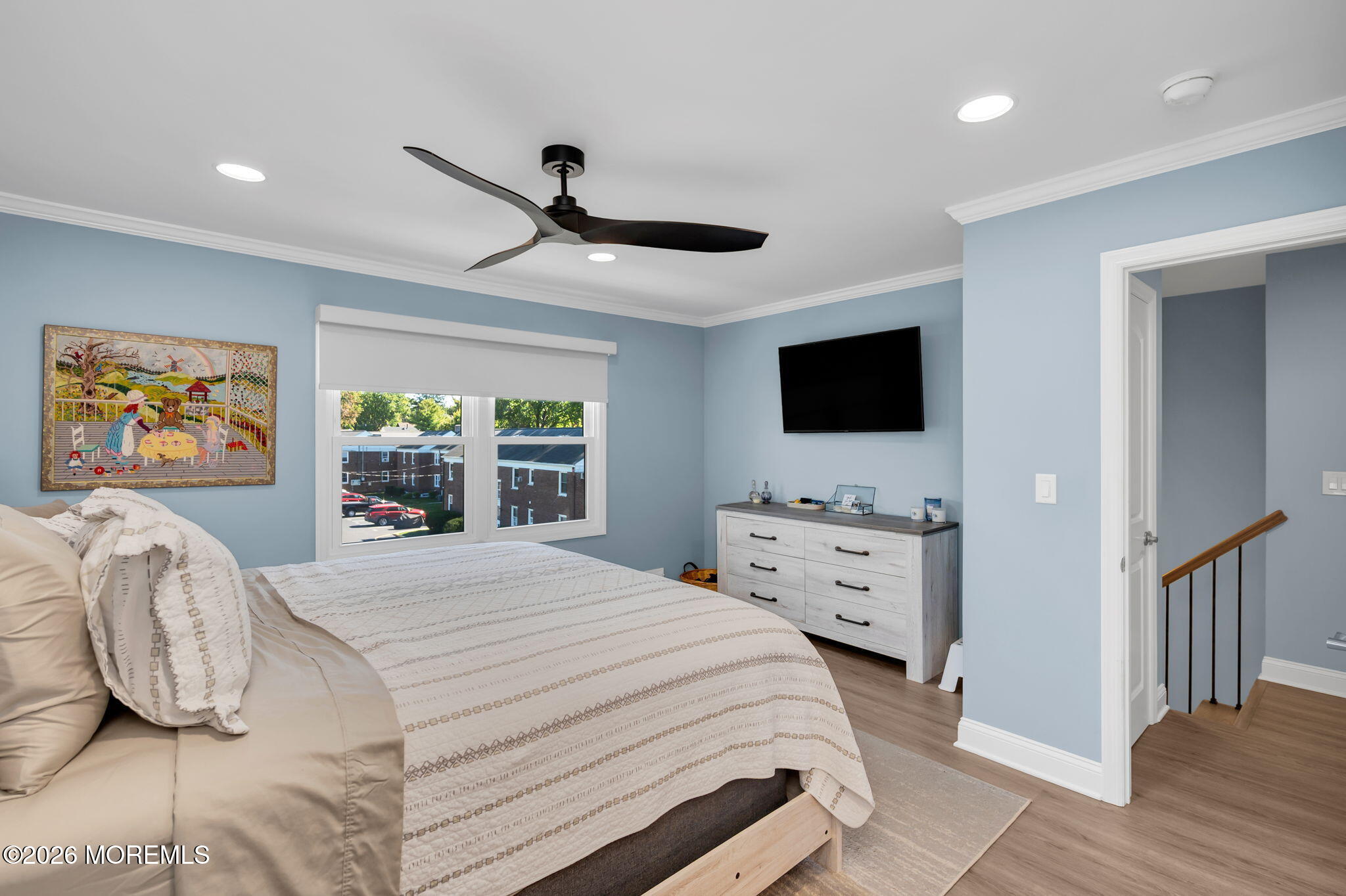 Bedroom with pale blue walls, a patterned bedspread, and windows overlooking nearby buildings.